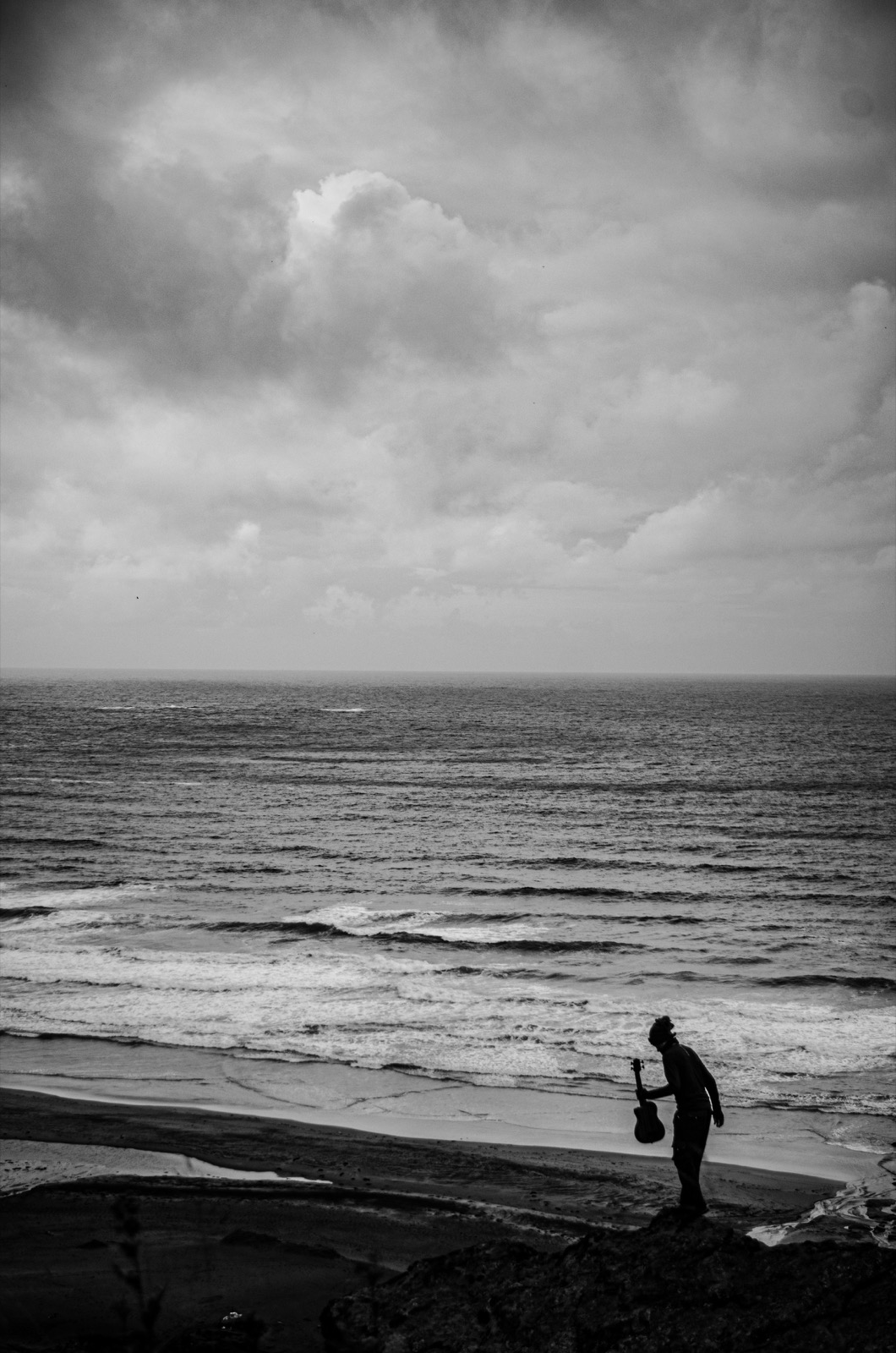 Marc Papillon, guitare à la main, sur une plage face à la mer sous un ciel chargé.